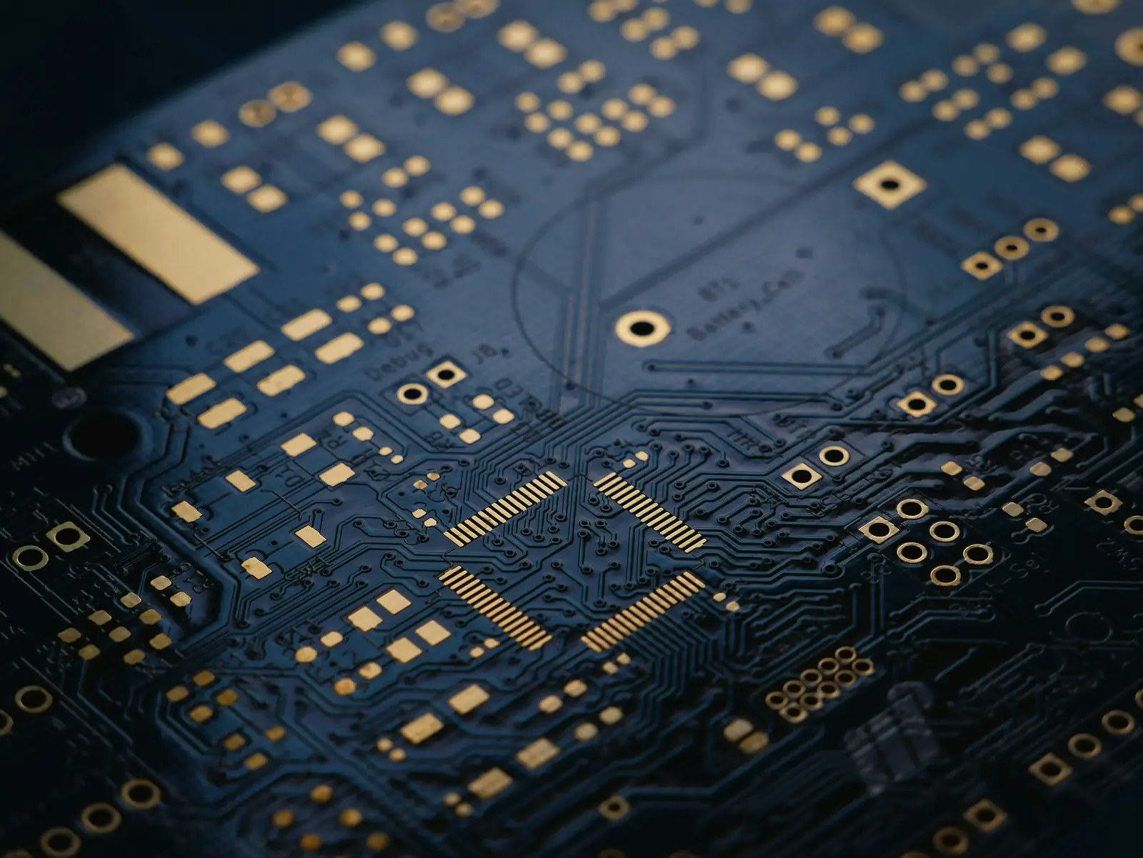 Macro shot of a dark-blue printed circuit board with gold traces and pads and a chip footprint in the centre, shallow depth of field