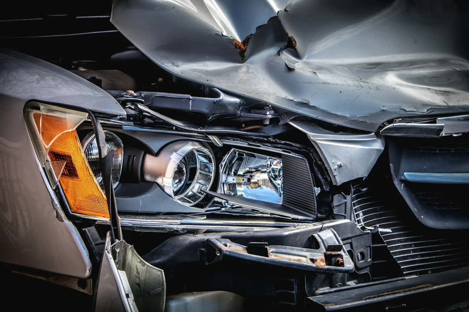 Gros plan sur l'avant accidenté d'une voiture grise, capot plié et optique de phare à nu