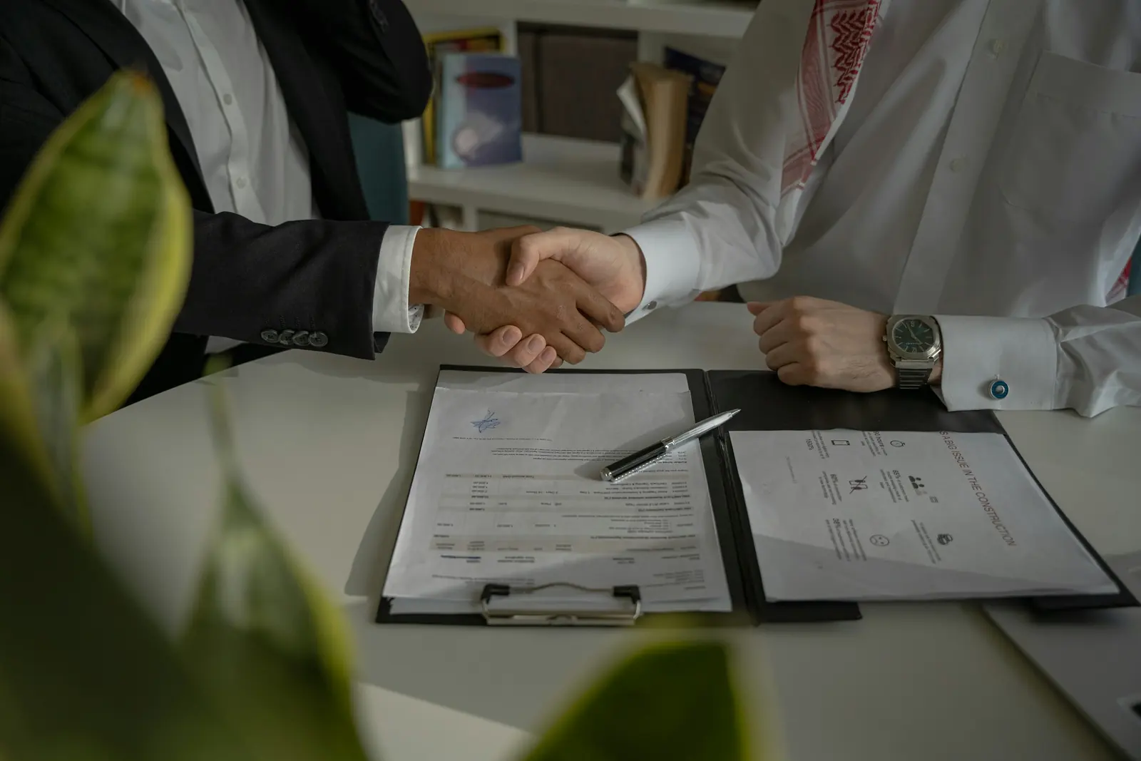 Poignée de main entre deux hommes d'affaires, l'un en costume et l'autre en tenue traditionnelle du Golfe, au-dessus de documents