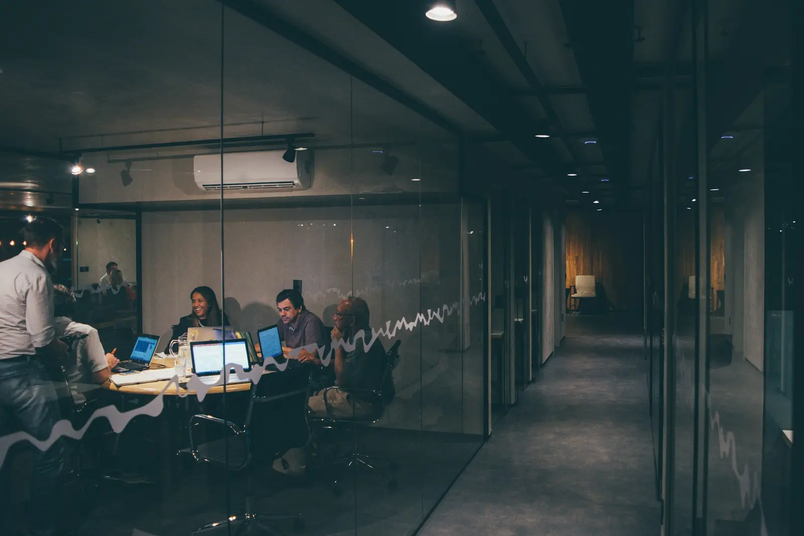Team meeting in the evening inside a glass-walled room of a modern office, with laptops open